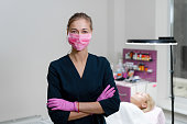 Cosmetology cabinet client lies on couch. Beautician stands in pink medical mask and smiles.