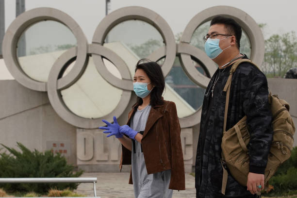 Couple wears a protective mask as they walks past the Olympics rings at the Olympic park on March 25, 2020 in Beijing, China. The 2020 Tokyo Olympics...