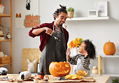 Ethnic father removing pulp from ripe pumpkin while carving jack o lantern with little son for Halloween