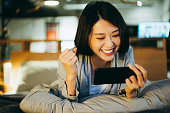 Excited young Asian woman lying on the bed in the bedroom, playing mobile game on smartphone in the evening at home