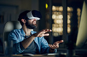 Excited young bearded man sitting in front of computer and gesturing hands while testing new app via VR device
