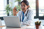 Female doctor waving and talking with colleagues through a video call with a laptop in the consultation.