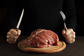 Female hands hold fork and knife and ready to eat raw meat on the wooden cutting board on dark background.