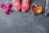 Fitness concept, pink sneakers, dumbbells, bottle of water and heart shaped plate with vegetables and berries on a grey background, top view, healthy lifestyle