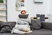 freezed young woman in warm clothes sitting on couch and hugging cushion at home