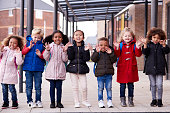 A group of smiling young multi-ethnic school kids wearing coats and carrying schoolbags standing in a row in walkway outside their infant school waving to camera, full length, front view