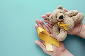 Hands holding children soft toy brown bear with yellow gold ribbon, Childhood cancer awareness