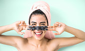 Happy smiling girl applying facial charcoal mask portrait - Young woman having skin care cleanser spa day - Healthy beauty clean treatment and cosmetology products concept - Aquamarine background