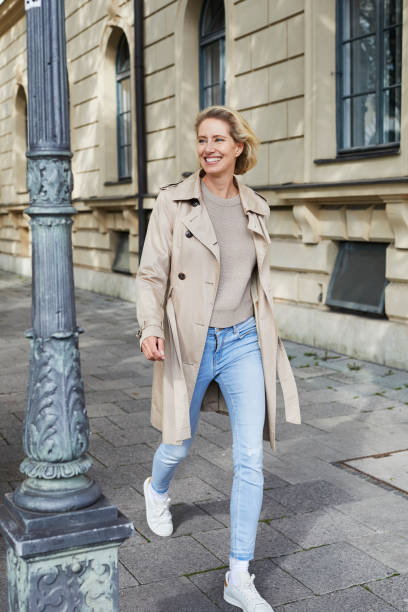 happy woman walking on pavement in the city - woman trench coat stock pictures, royalty-free photos & images