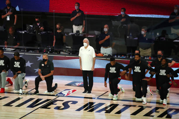 Head coach Gregg Popovich of the San Antonio Spurs wears a mask while players kneel before a NBA basketball game against the Houston Rockets at The...