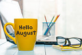 Hello August - inscription at yellow morning coffee or tea cup at business office background. Summer month, Calendar concept