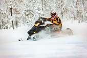Human in the outfit of a racer in a multi-color jumpsuit and a helmet, driving a snowmobile by the deep snow surface on background of snowy forest.
