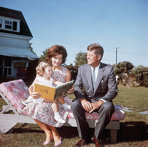 Jacqueline Kennedy and her husband Senator John F. Kennedy sit on a lounge chair at their summer home. Jacqueline reads to her daughter Caroline from...