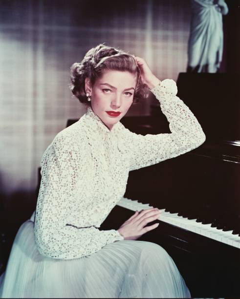 Lauren Bacall, US actress, wearing a white macrame blouse and a light blue skirt while posing beside a piano, with one hand on the keys, circa 1955.