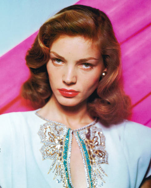 Lauren Bacall, US actress, wearing a white top with a keyhole necklace with sequinned decoration in a studio portrait, against a pink background,...