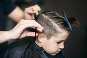 Little Boy Getting Haircut By Barber While Sitting In Chair At Barbershop.