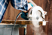Male carpenter using electric circular saw in home workshop with wood chips flying