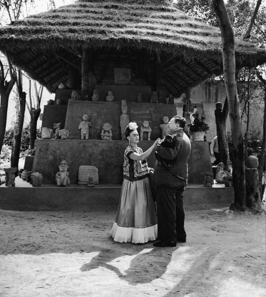 Married Mexican painters Frida Kahlo and Diego Rivera stand together with a pet monkey in front of thatchted-roof hut which houses a number of...