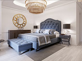 Modern bed in classic blue style with bedside table and lamp. Large glass chandelier over. A dresser with a decor and a golden mirror above. Modern bedroom.
