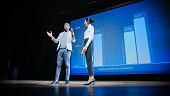 On Stage, Successful Female CEO and Male COO Speakers Present Company's New Product, Show Infographics, Statistics on Big Screen, Talk About Growth. Live Event, Tech Startup, Business Conference
