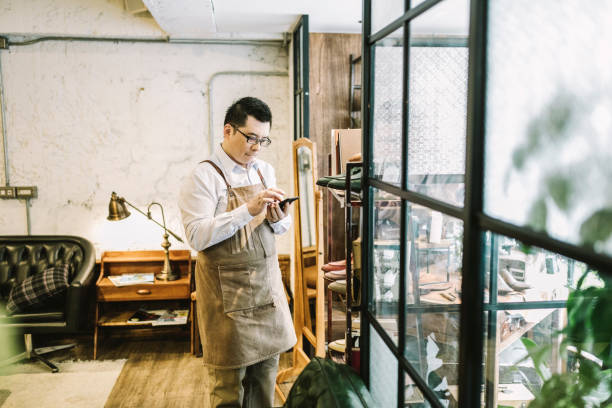 Small business owner sending voice message while standing by window. Entrepreneur using smart phone while looking away. He wearing apron in store.