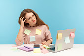 Pensive upset woman employee in nerd eyeglasses sitting at workplace office, all covered with sticky notes