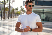 Portrait of handsome young man wearing sunglasses and white tshirt, posing on city street background. Casual style.