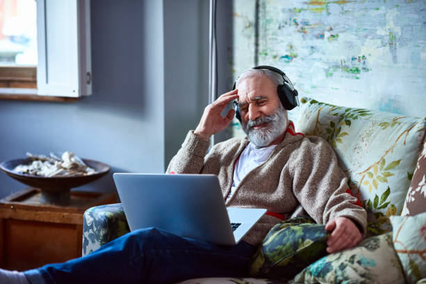 portrait of mature man streaming movie on laptop wearing headphones - streaming - fotografias e filmes do acervo