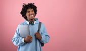 Positive African American teen student with backpack, tablet pc and headphones studying online on pink background