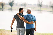 Rear view of athletic father and son talking while walking embraced by the lake.