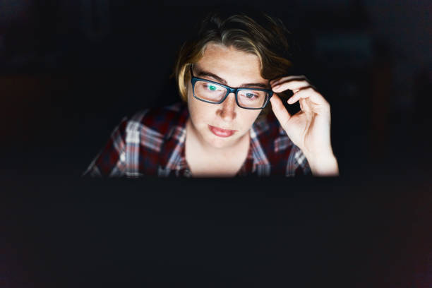 serious young woman, lit by monitor against black, adjusts spectacles - fraud job stock pictures, royalty-free photos & images