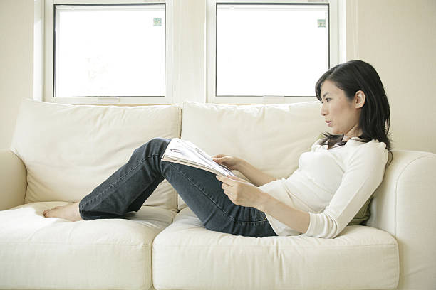 Side profile of a mid adult woman reclining on a couch and holding a magazine
