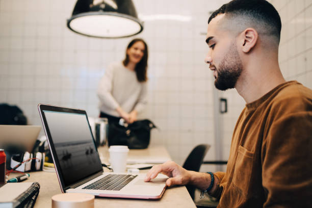 side view of young male hacker using laptop while female colleague working at small office - professional software developer stock pictures, royalty-free photos & images