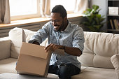 Smiling african man customer opening cardboard box parcel on sofa