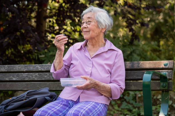 smiling senior asian woman relaxing in park and eating blueberries - old asian woman eating alone stock pictures, royalty-free photos & images