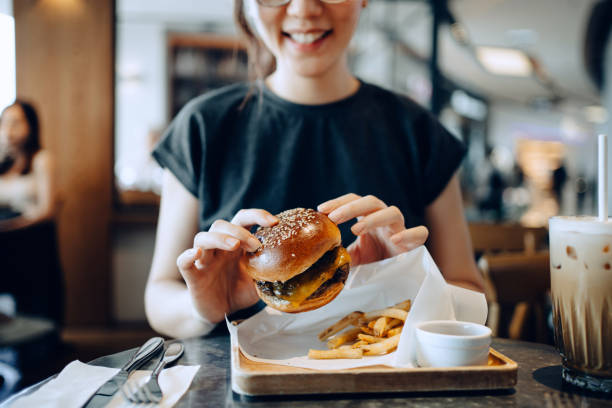 smiling young asian woman enjoying freshly made delicious burger with fries and a glass of iced coffee in a cafe - ハンバーグ ストックフォトと画像