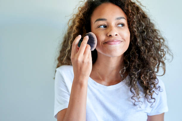 Smiling young woman applying face powder with make-up brush against white wall