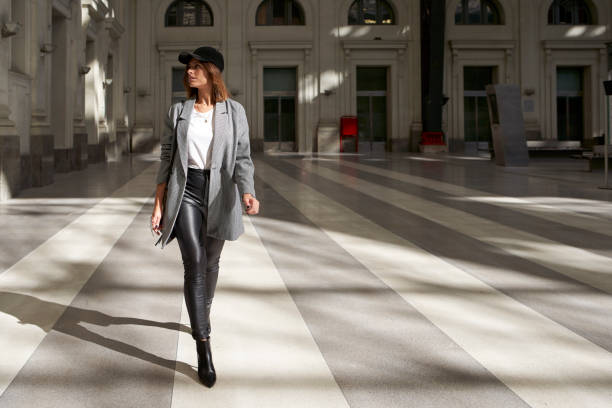 stylish woman wearing white t-shirt, long blazer, black leather pants and black cap walking alone through a public corridor. - blazer stock pictures, royalty-free photos & images