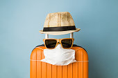 Suitcase with hat, sunglasses and protective medical mask on pastel blue background minimal creative coronavirus covid-19 travel concept.