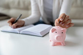 Thrifty woman writing daily expenses put coin into piggy bank