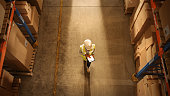 Top-Down View: Worker Wearing Hard Hat Checks Stock and Inventory Using Digital Tablet Computer in the Retail Warehouse full of Shelves with Goods. Working in Logistics, Distribution