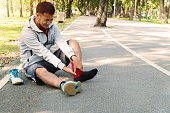 Unhappy young man runner knee injury and pain while sitting on the road in the park. Male athlete suffering from painful knee with accident while running in public park. Injury from workout concept.