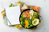 Vegan healthy balanced diet Vegetarian buddha bowl with blank notebook and measuring tape. hickpeas, broccoli, pepper, tomato, spinach, arugula and avocado in plate on white background. Top view