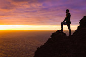 Visionary man standing on top of cliff edge staring at colorful sunset by the sea in Gran Canaria