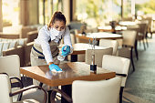 Waitress with a face mask and gloves cleaning tables with disinfectant in a cafe.