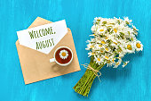Welcome August text on white card in craft envelope, bouquet white chamomiles flowers and cup of tea on blue background. Concept Hello august. Greeting card Flat Lay