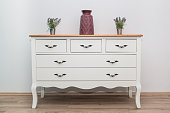 White wooden dresser with three vases and flowers on white wall background. Chest of drawers close up.