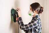 Woman in protective mask working with electric sander to smooth plaster wall surface, room renovation concept