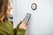 Woman regulating heating temperature with phone and thermostat at home