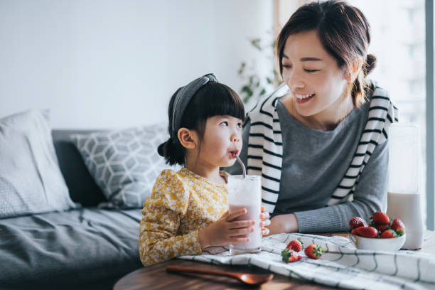 young asian mother making healthy homemade strawberry smoothie with lovely little daughter at home. mother and daughter smiling at each other joyfully and enjoying the drink. healthy eating and healthy lifestyle - オーツミルク ストックフォトと画像